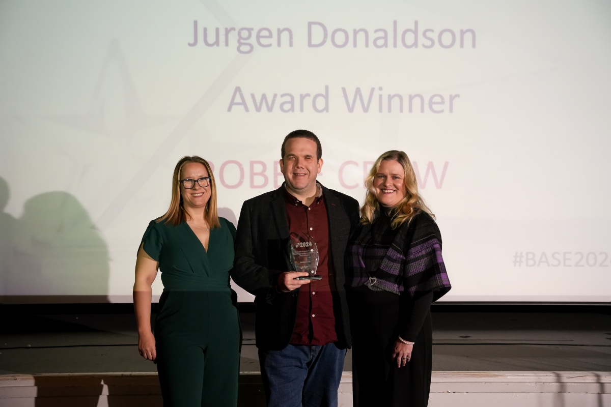 Robbie Crow is smiling, standing on a stage at an awards presentation evening, holding an engraved glass award in his hand. Robbie is a white male with a blazer and maroon shirt on. He is stood with Laura Davis on his left is a white woman with blonde, straight, shoulder length hair , wearing a green jumpsuit, she’s smiling and has her arm around Robbie. On the right of Robbie, also with her arm around him ,is Kerrie Moncrieff , a woman with blonde shoulder-length wavy hair , wearing a plaid -patterned top and she’s smiling