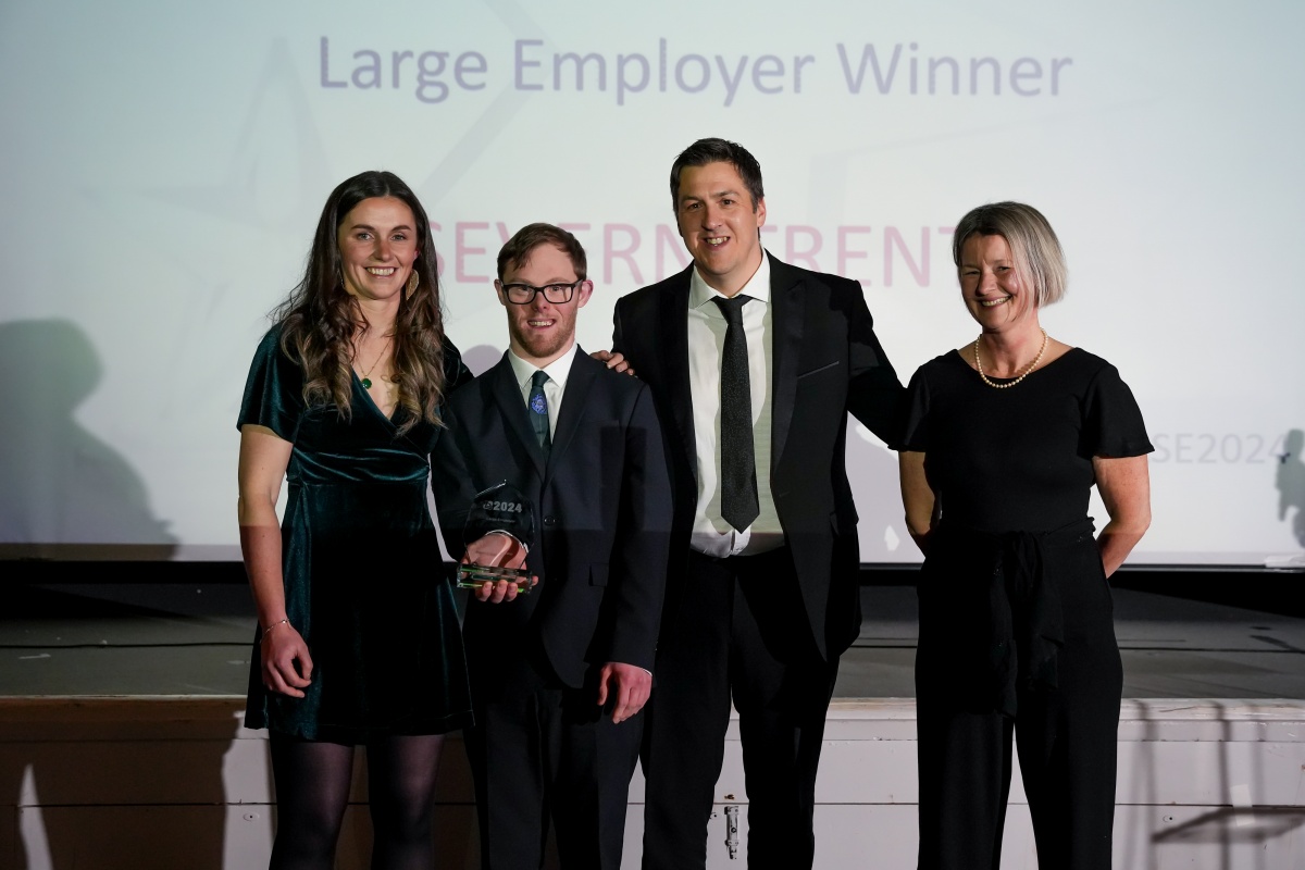 Left is a young woman wearing a green, velvet dress with long brown hair, she is smiling and has her arm on the shoulder of the man next to her. He has short brown hair, he is wearing glasses and a navy blue suit . He is holding up a glass award. Next is a tall white man, with dark short hair, wearing a suit. He is smiling. On the far right is a white woman with jaw-length blonde hair, she is wearing a black jumpsuit  and a smile  Next to him is  and tie har
