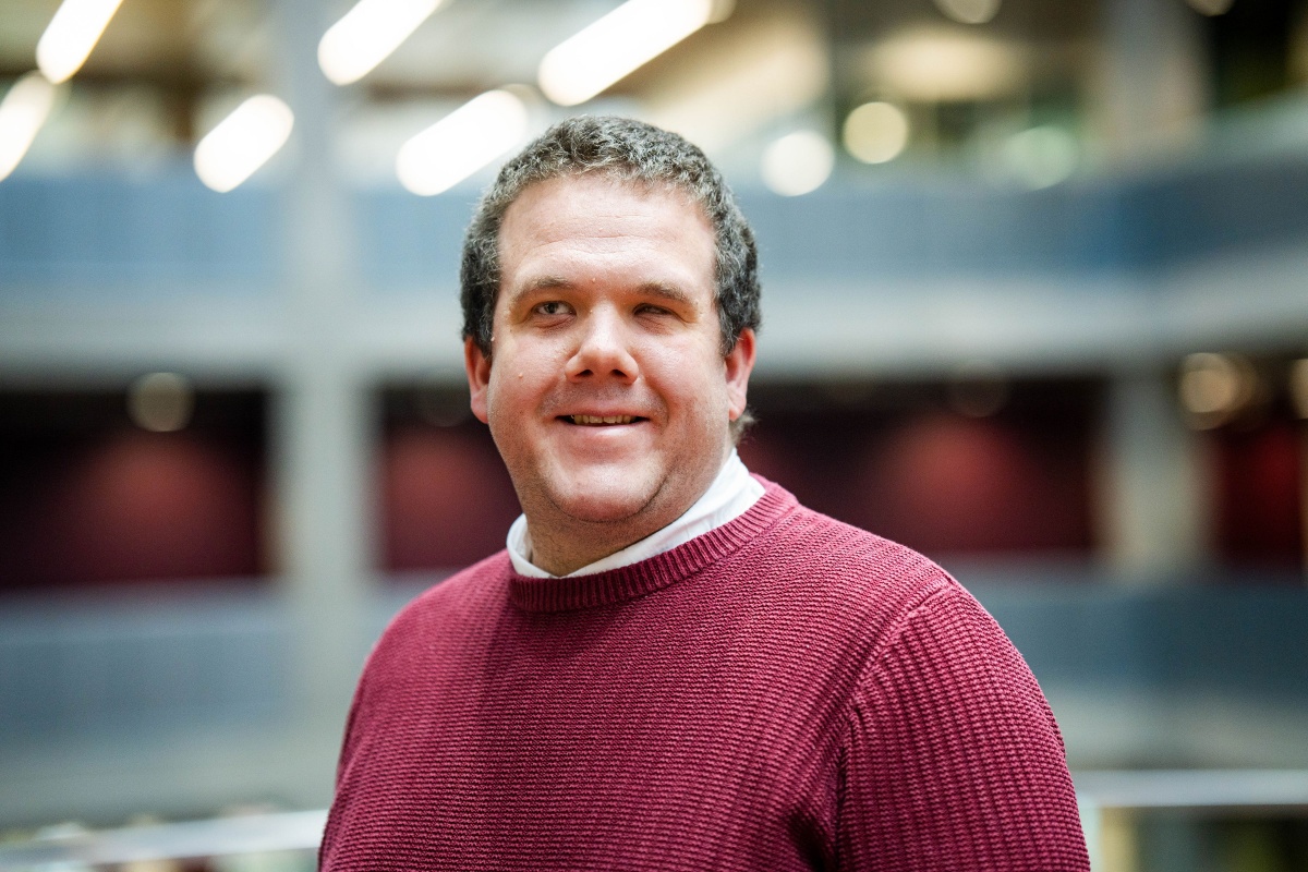 Robbie, a white male with short hair, stands in the foreground with a blurry office-type scene in the background. Robbie is smiling slightly at the camera and wearing a maroon jumper with a white collar peeking through. Only his head and shoulders are in view.